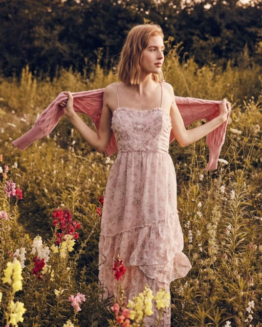 Woman in floral dress stands in flower field with pink cardigan, looking left in medium shot. Golden light, outdoor setting.
