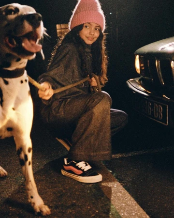 Young woman in a pink beanie and denim jumpsuit walking a Dalmatian dog at night near a car by Manu Fauque