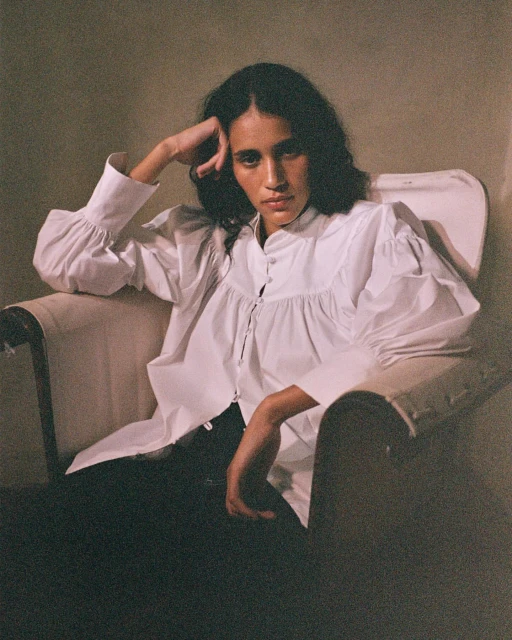 Elegant woman with dark hair in a white blouse poses sitting in a chair against a neutral background in a fashion portrait.