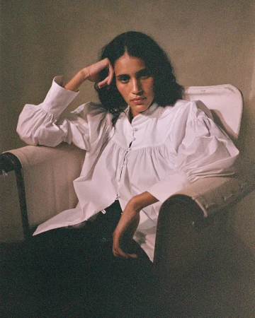 Elegant woman with dark hair in a white blouse poses sitting in a chair against a neutral background in a fashion portrait.