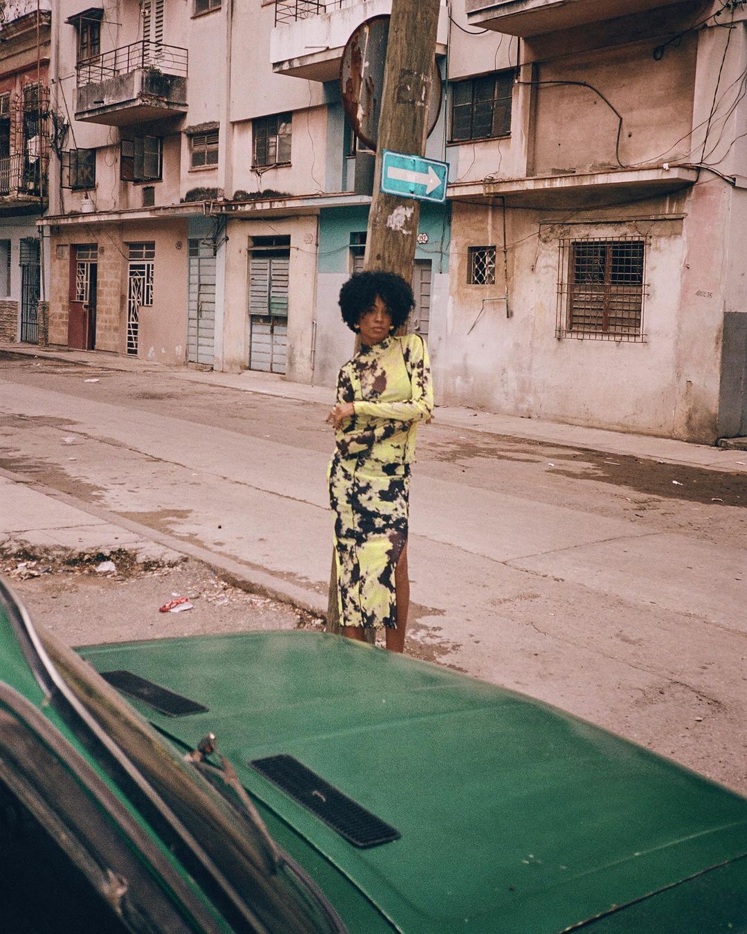 An adult woman in a patterned dress stands on a Havana street next to a green vintage car, with weathered buildings and a directional sign.