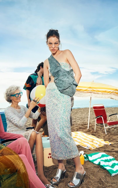 A woman in a sequin dress poses on a beach with a casual bag, with other people, chairs, and beach accessories visible in the background.