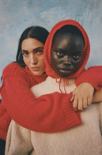 Two women embrace in a studio portrait. One woman with dark hair wears a red knit sweater, while the other wears a cream-colored hoodie.