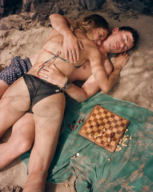 A couple lounges on a sandy beach next to a chessboard. The woman embraces the man as they relax on a green towel by the water.