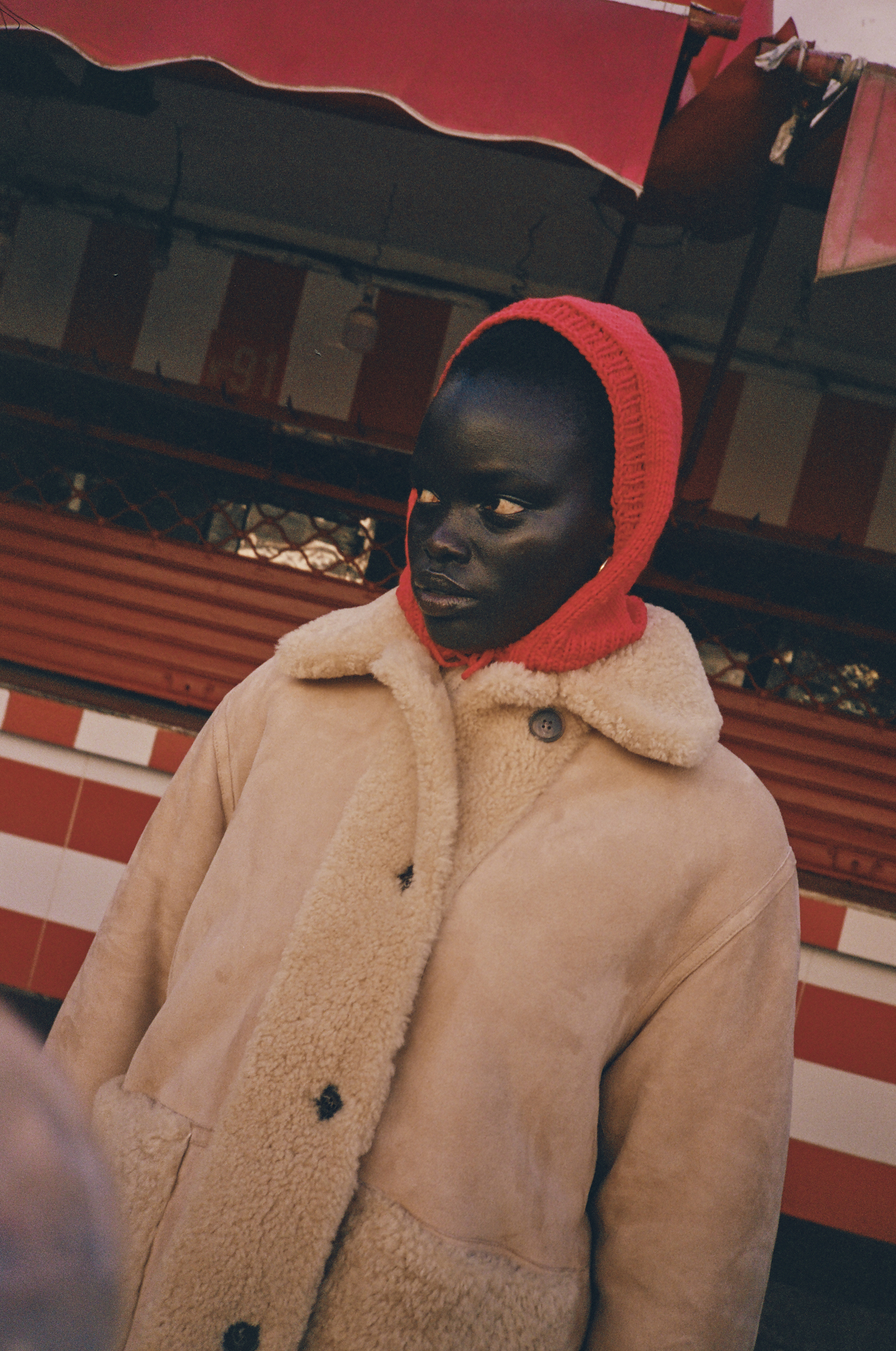 Adult female wearing a beige shearling coat and a red knitted balaclava stands against a red striped building outdoors.