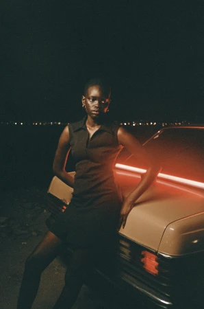 A dark-skinned woman leaning against a vintage car at night, illuminated by red neon light under a dark sky.