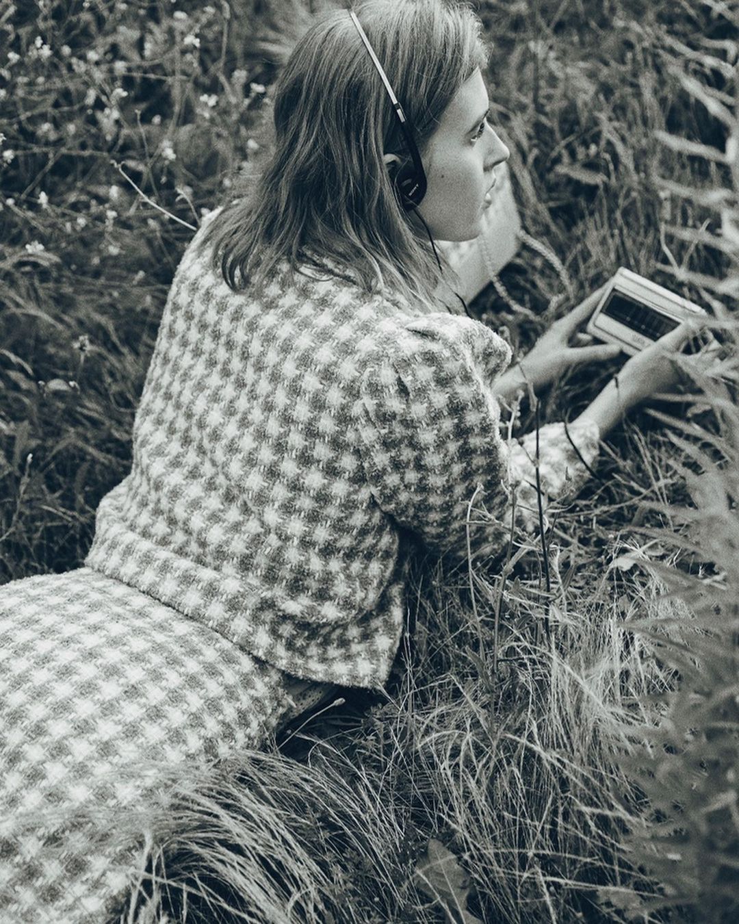 Woman in patterned jacket and skirt lies in tall grass, listening to a radio with headphones, in a vintage black and white aesthetic.