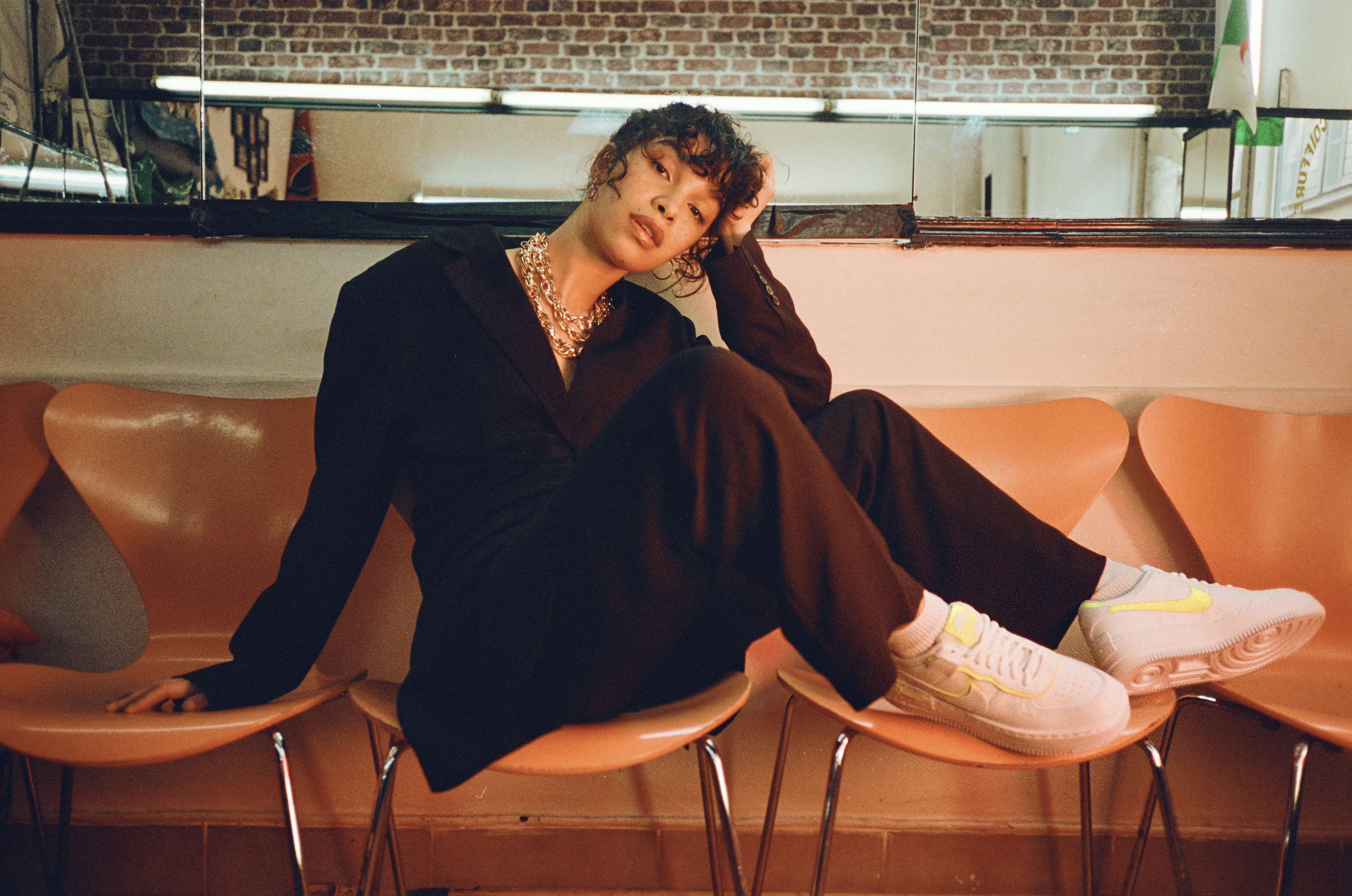 A young adult woman in a black suit sits on an orange chair, legs crossed, in a room with a brick wall and a large mirror. 