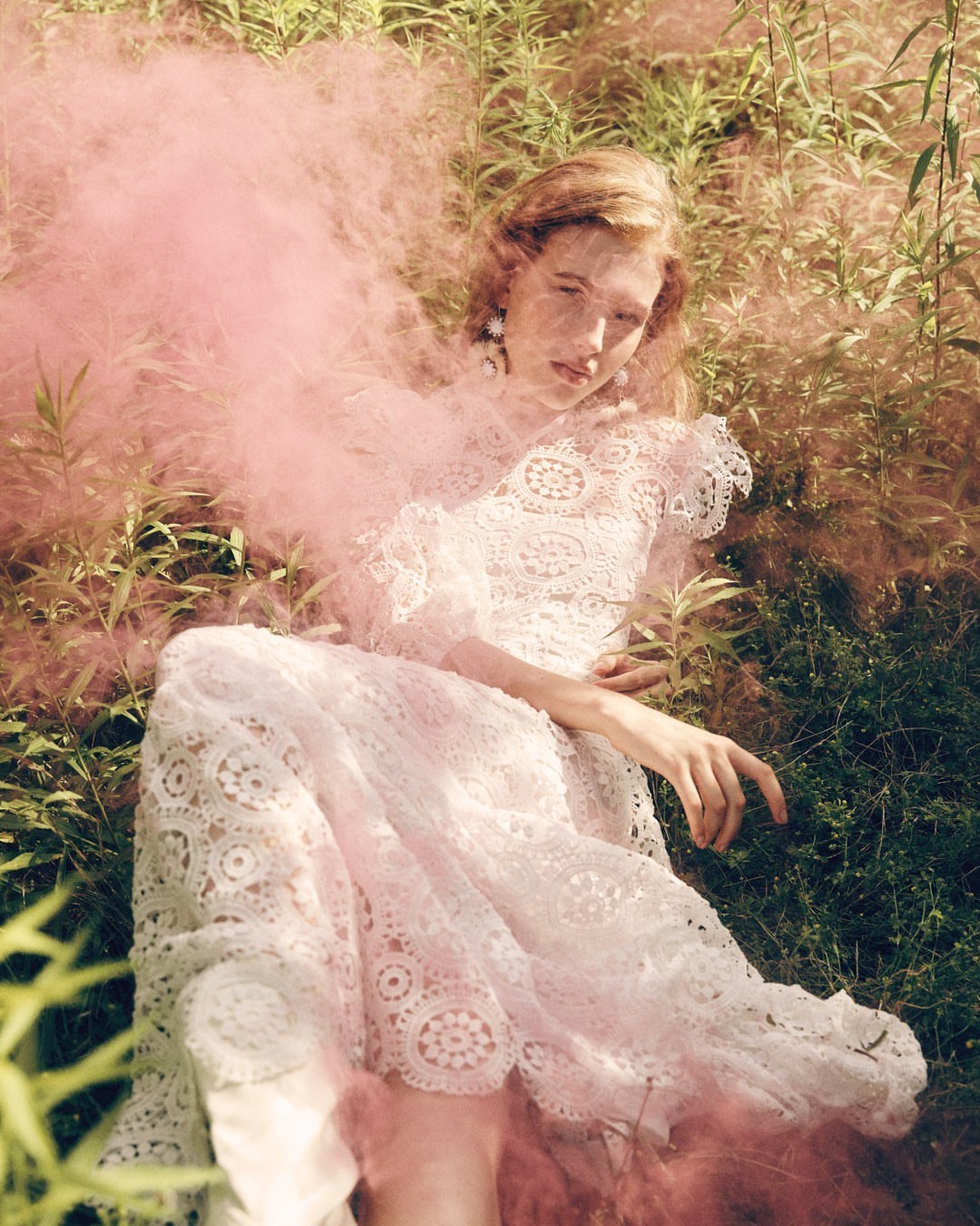 Woman in a lace dress lies in a field of tall grass with pink smoke or fog surrounding her in a soft, ethereal fashion shot.