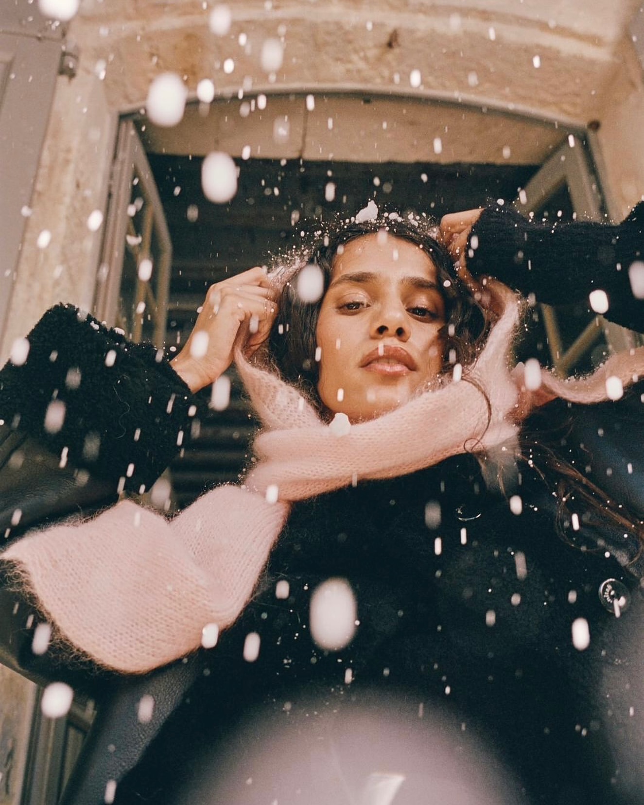 Young woman in black coat and pink scarf poses in a snowy outdoor setting with a neutral stone wall and door frame behind her.