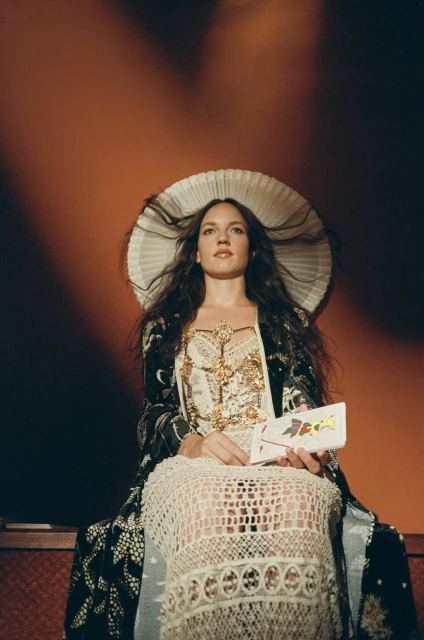 A woman in a decorative white dress and headpiece sits against an orange background, holding a small white box in her hands.