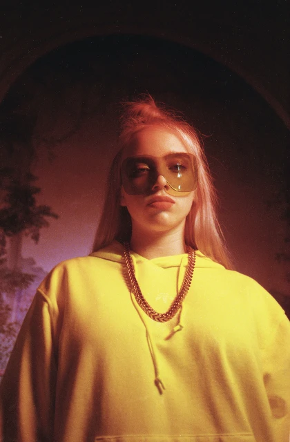 Portrait of a young adult woman with blond hair, wearing yellow hoodie, and sunglasses, posing in an indoor studio with mood lighting.