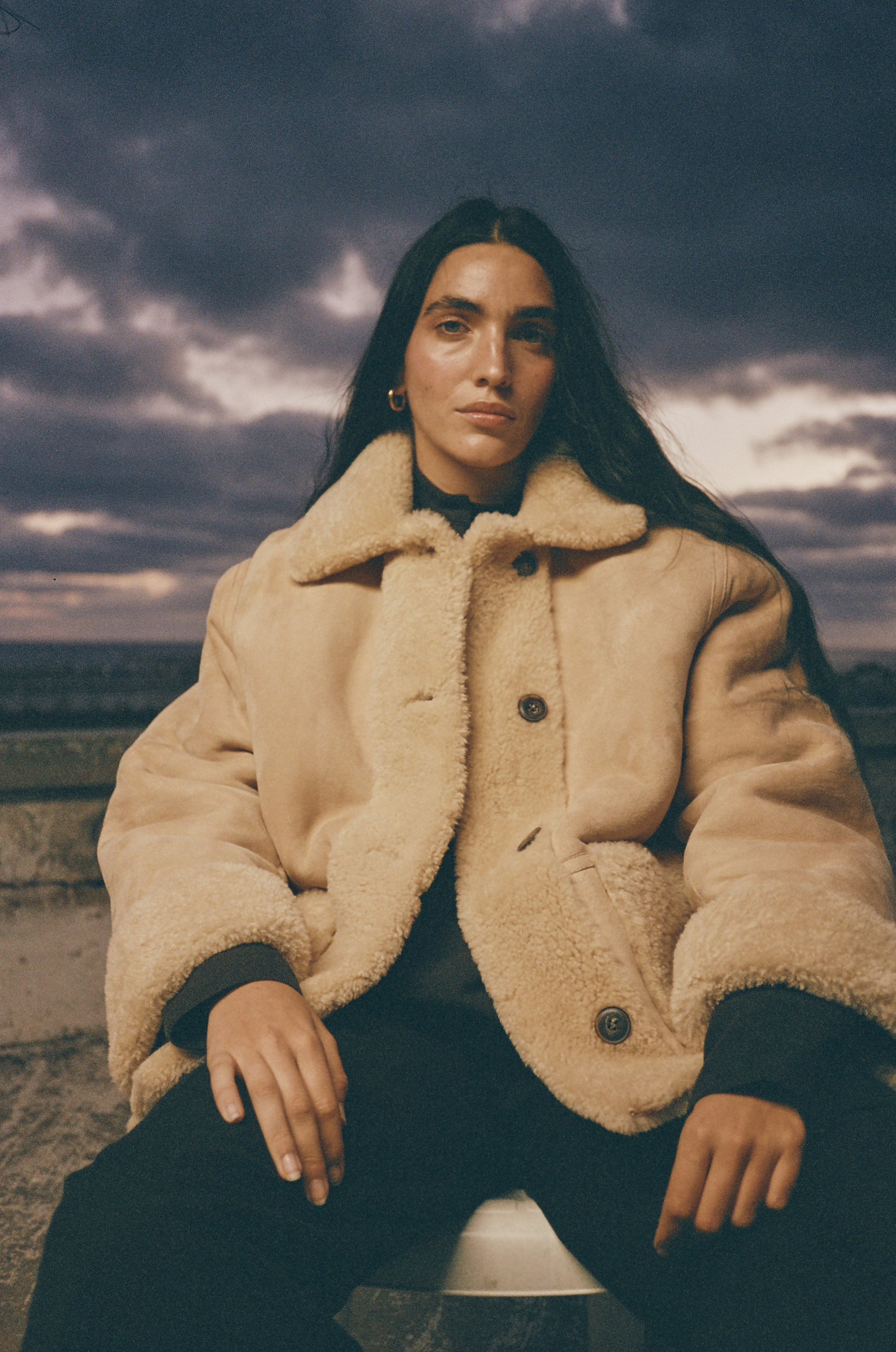 Brunette woman poses outdoors in a tan shearling coat, black pants, and gold earrings, against a dark sky and landscape backdrop.