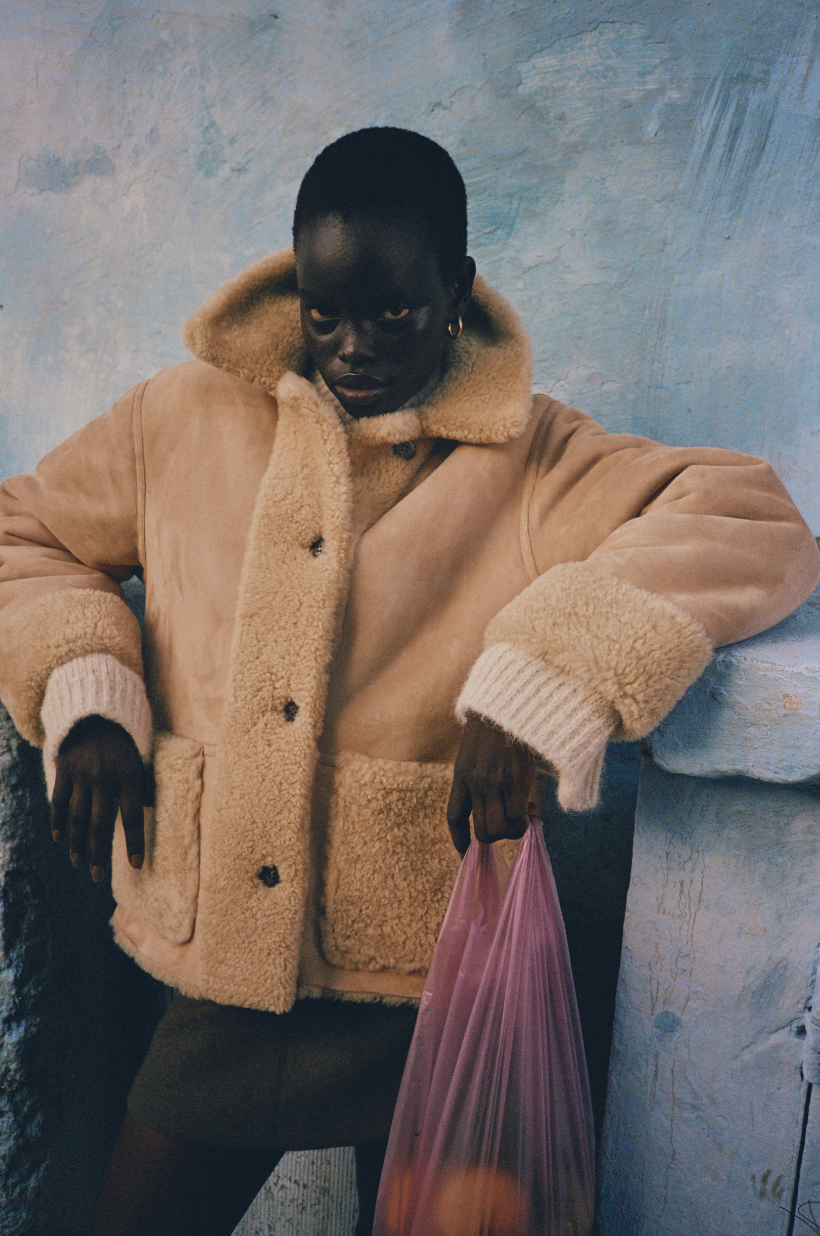 Dark-skinned model in a cream-colored coat poses against a blue wall, holding a pink bag filled with oranges in a medium indoor shot.