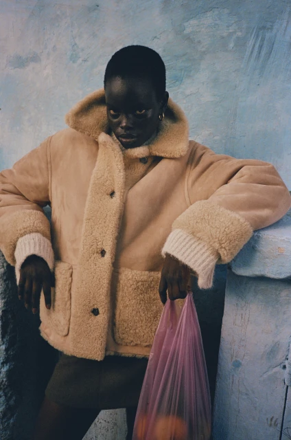 Dark-skinned model in a cream-colored coat poses against a blue wall, holding a pink bag filled with oranges in a medium indoor shot.