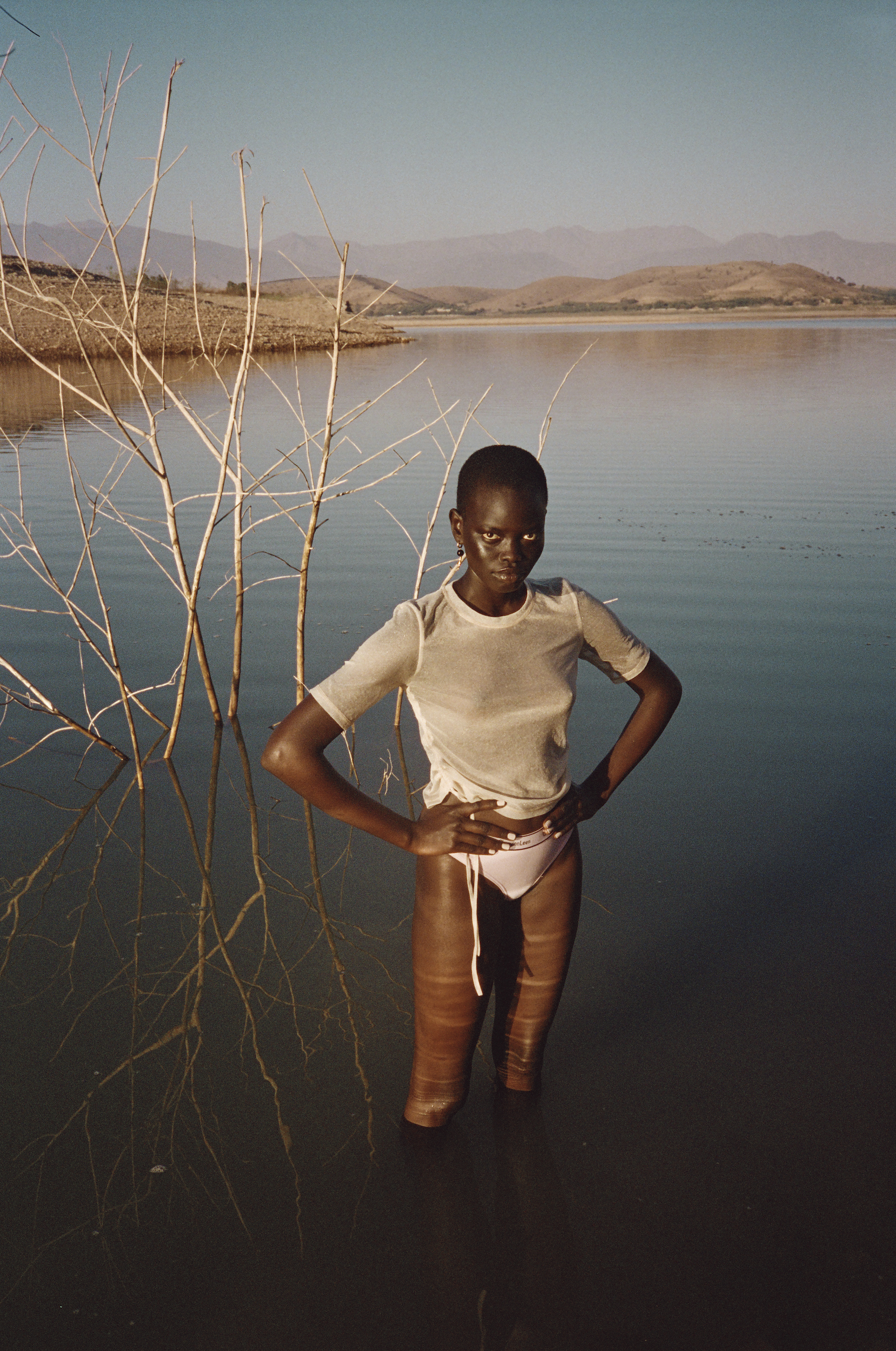 A solitary figure, an African woman stands waist-deep in tranquil water, surrounded by delicate bare tree branches, under a clear sky.