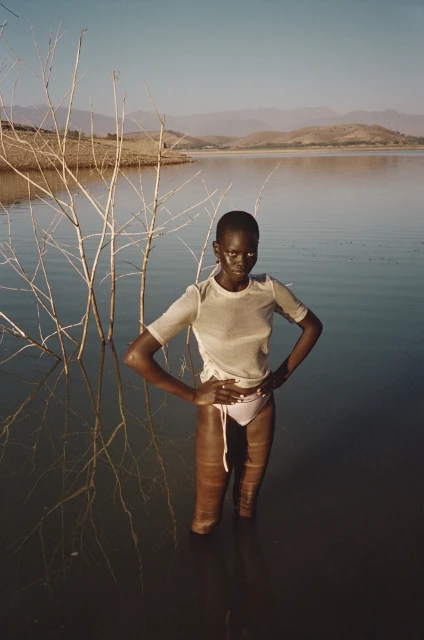 A solitary figure, an African woman stands waist-deep in tranquil water, surrounded by delicate bare tree branches, under a clear sky.