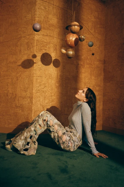 Woman in patterned pants looks up at a hanging mobile of planet-like orbs in an indoor studio with wood-paneled walls and green carpet.