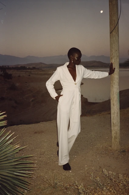 A dark-skinned woman poses in a white jumpsuit near a utility pole on a hillside at dusk, with mountains visible in the background.