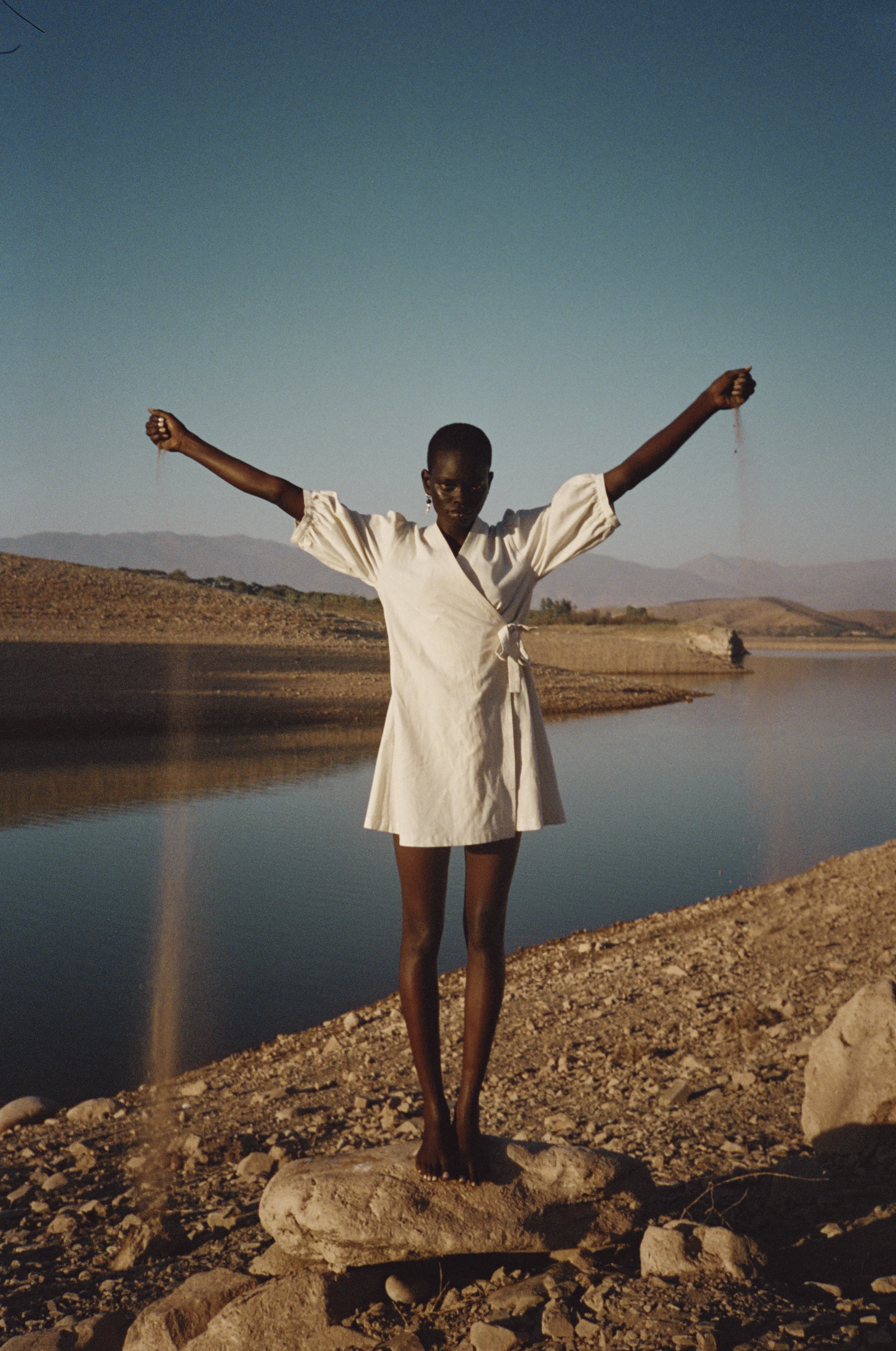 A slender, dark-skinned woman in a light dress stands on a rock with her arms outstretched against a lake and mountain backdrop.