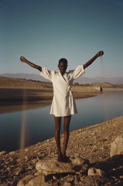 A slender, dark-skinned woman in a light dress stands on a rock with her arms outstretched against a lake and mountain backdrop.