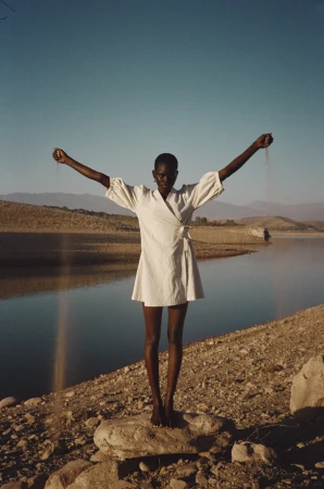 A slender, dark-skinned woman in a light dress stands on a rock with her arms outstretched against a lake and mountain backdrop.