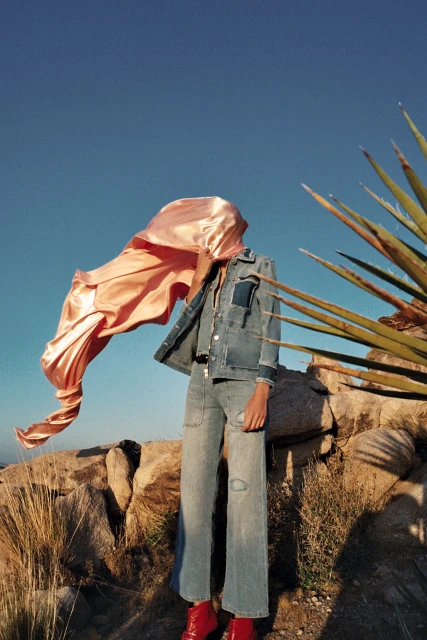 A person wearing a denim jacket and jeans with a peach-colored silk scarf blowing in the wind stands on a rocky desert landscape under a blue sky.