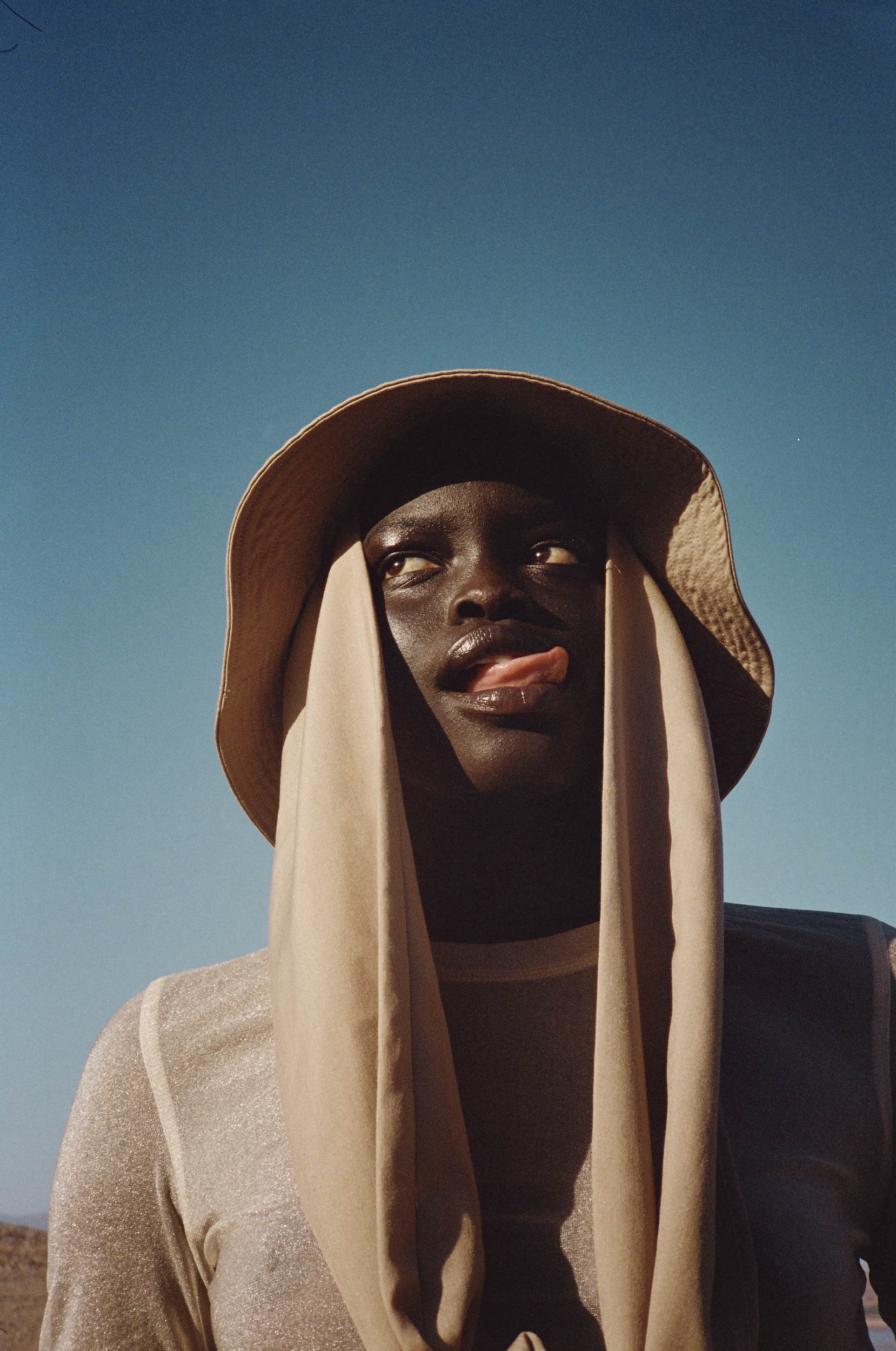 Portrait of a young adult with dark skin in a sun hat and sheer draped top, sticking out tongue, against a clear blue sky.