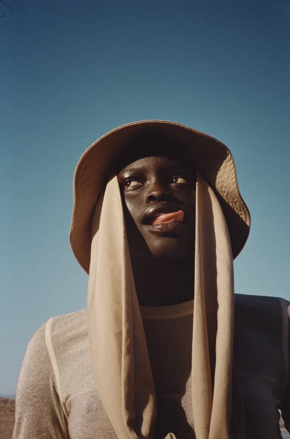 Portrait of a young adult with dark skin in a sun hat and sheer draped top, sticking out tongue, against a clear blue sky.