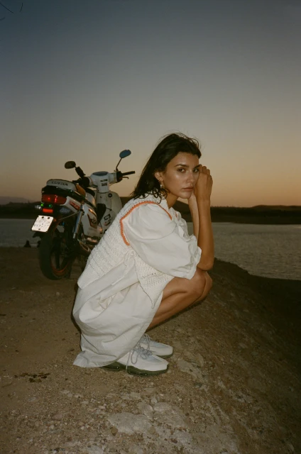 A brunette woman in a white dress and sneakers poses near a scooter by the water at dusk, featuring soft, natural lighting and a tranquil vibe.