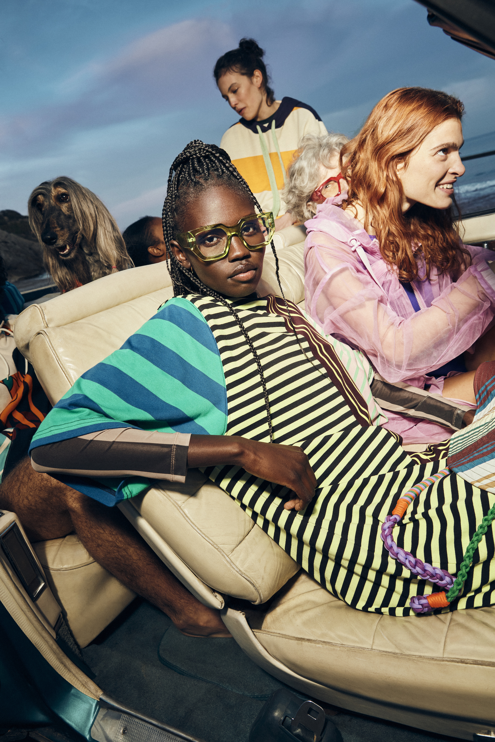 Group of people and a dog in a vintage car; adult women with bright, retro-inspired fashion in outdoor setting with sunset light.