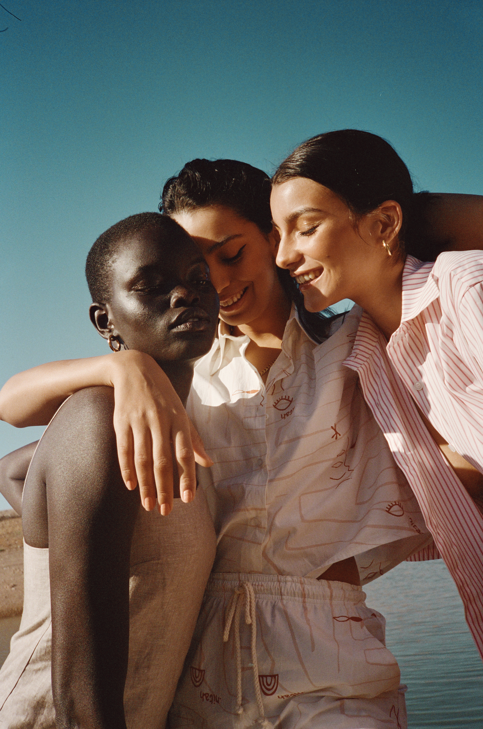 Three women embracing in matching casual outfits. The photo features a diverse trio against a clear sky and the edge of a body of water.
