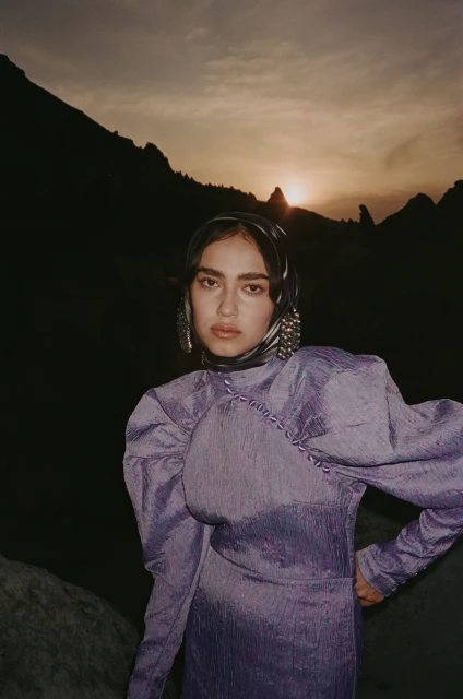 A fashionable woman in a lavender dress and headscarf poses against a dramatic mountain backdrop at sunset, creating a striking visual contrast.