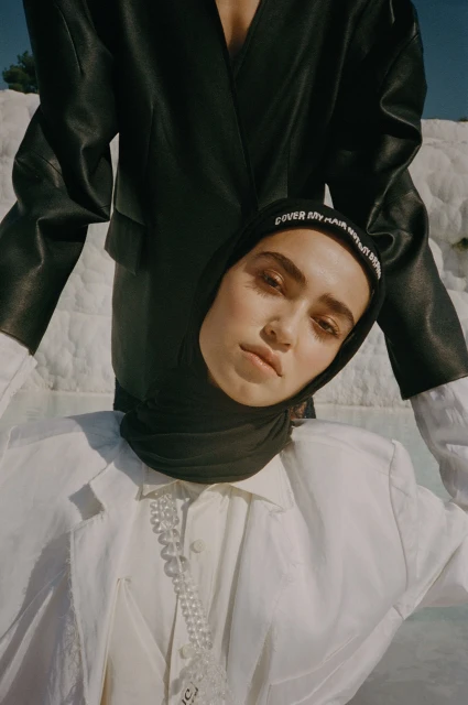 Fashion editorial featuring a woman in a black headscarf and a white blazer, styled with a pearl necklace against a travertine terrace backdrop.