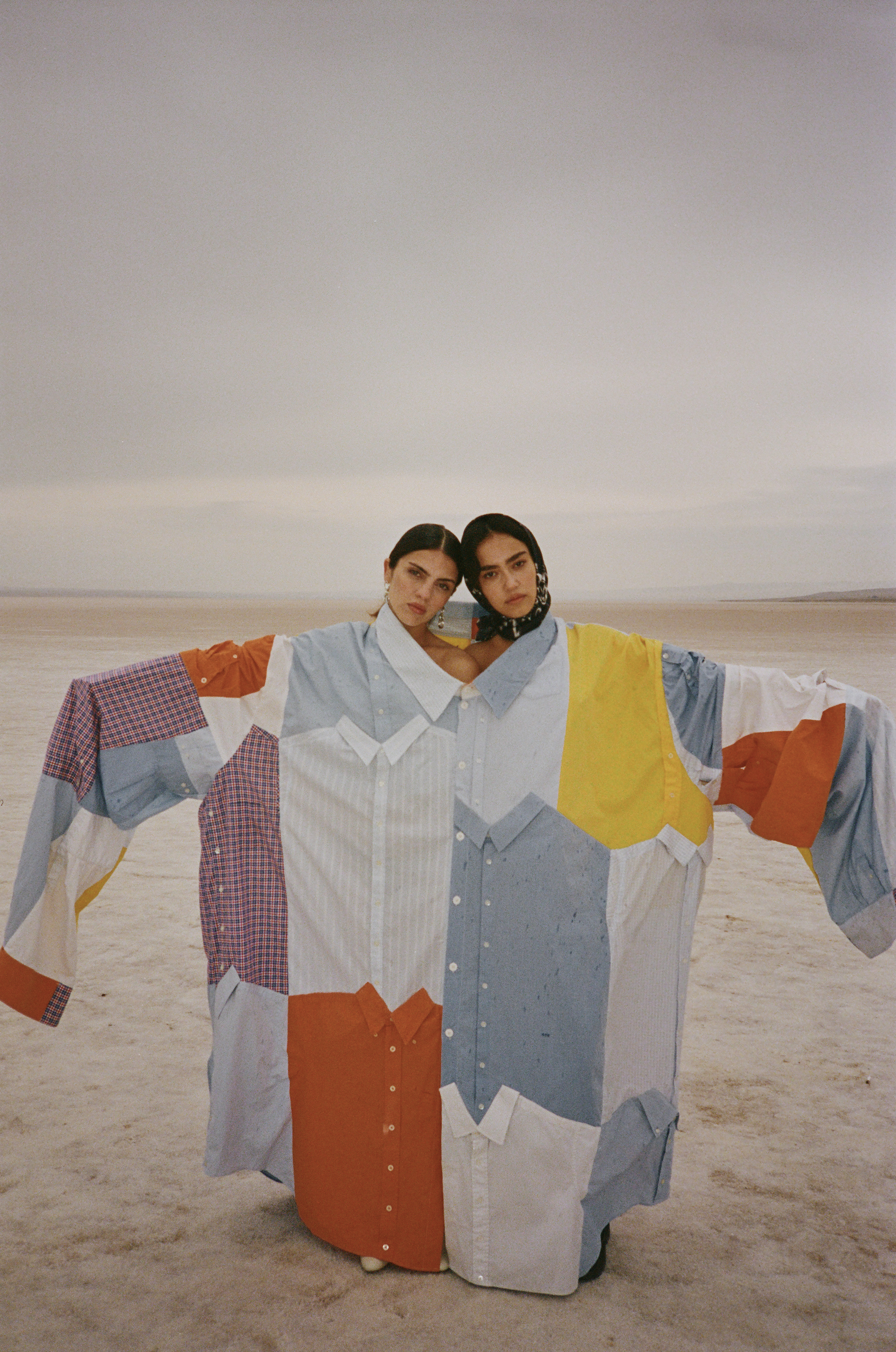 Two adult females in a field, wearing a single oversized patchwork shirt made of repurposed collared shirts, posing with outstretched arms.