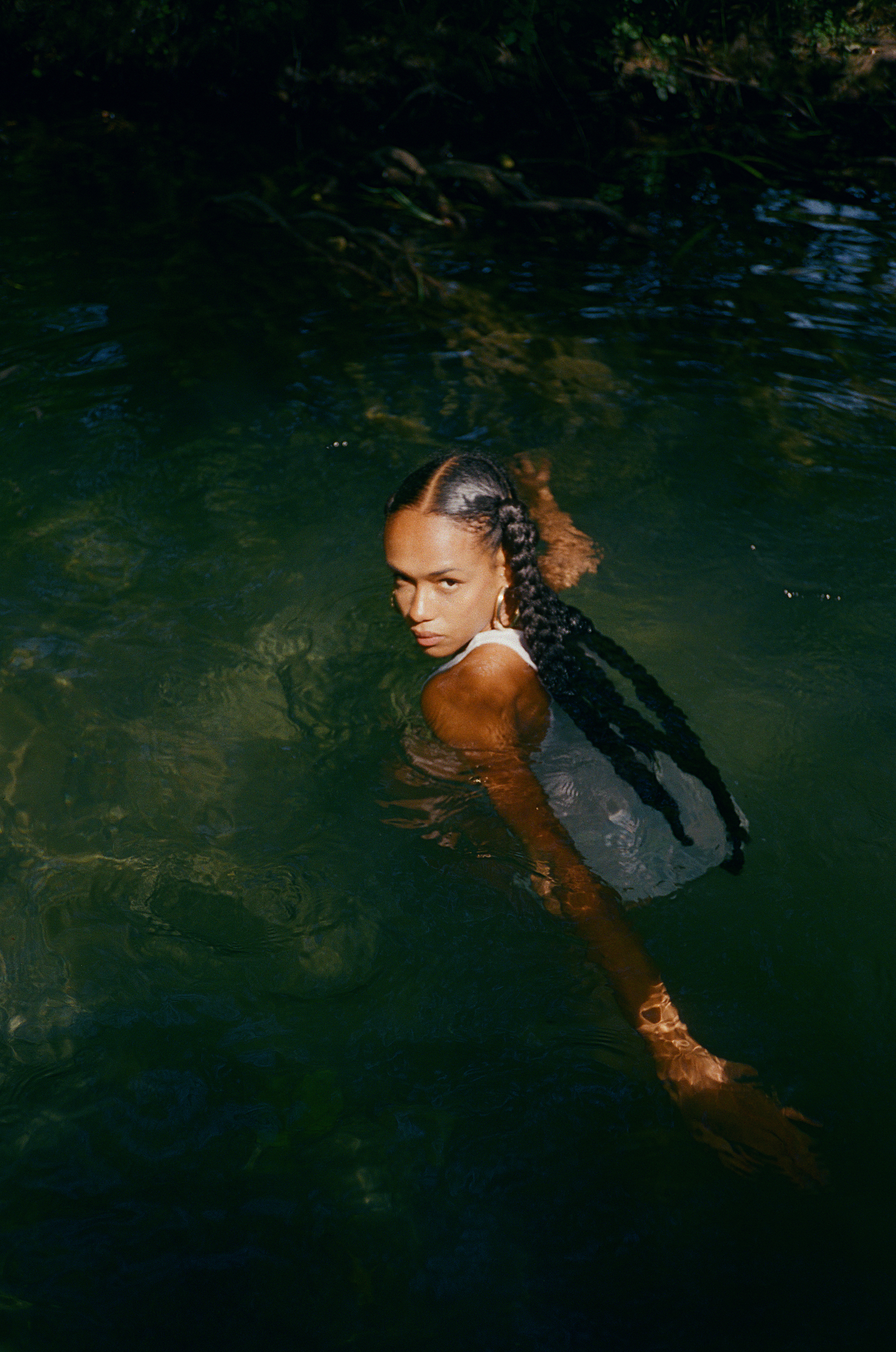 Woman in a white tank top, partially submerged in water, looking at the camera with a neutral expression, with dark hair.