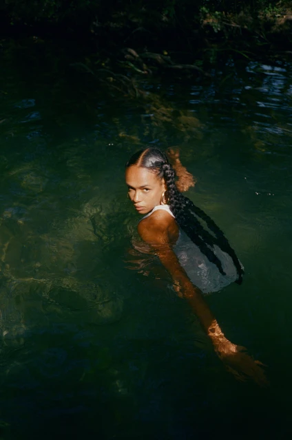 Woman in a white tank top, partially submerged in water, looking at the camera with a neutral expression, with dark hair.