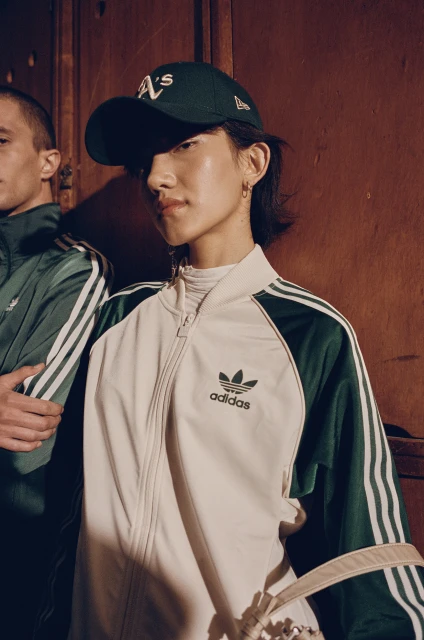 Two people wearing athletic apparel, one in a baseball cap and Adidas track jacket, against a dark wooden background.