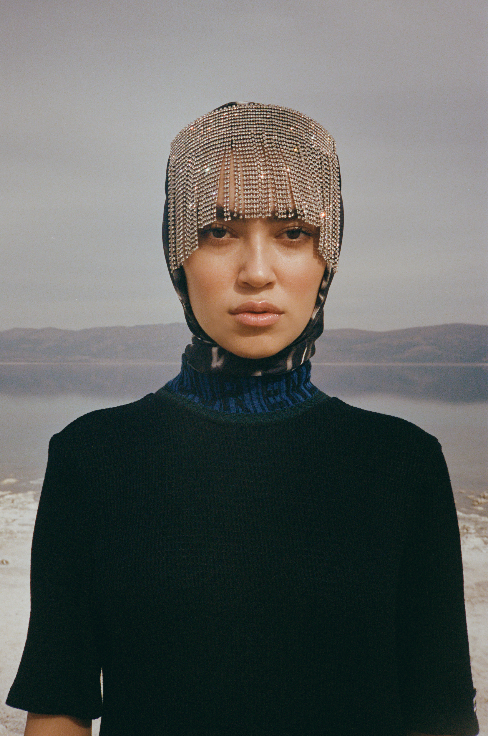 Woman in a black top and jeweled head covering poses outdoors with a neutral expression. The background features a lake and distant mountains.