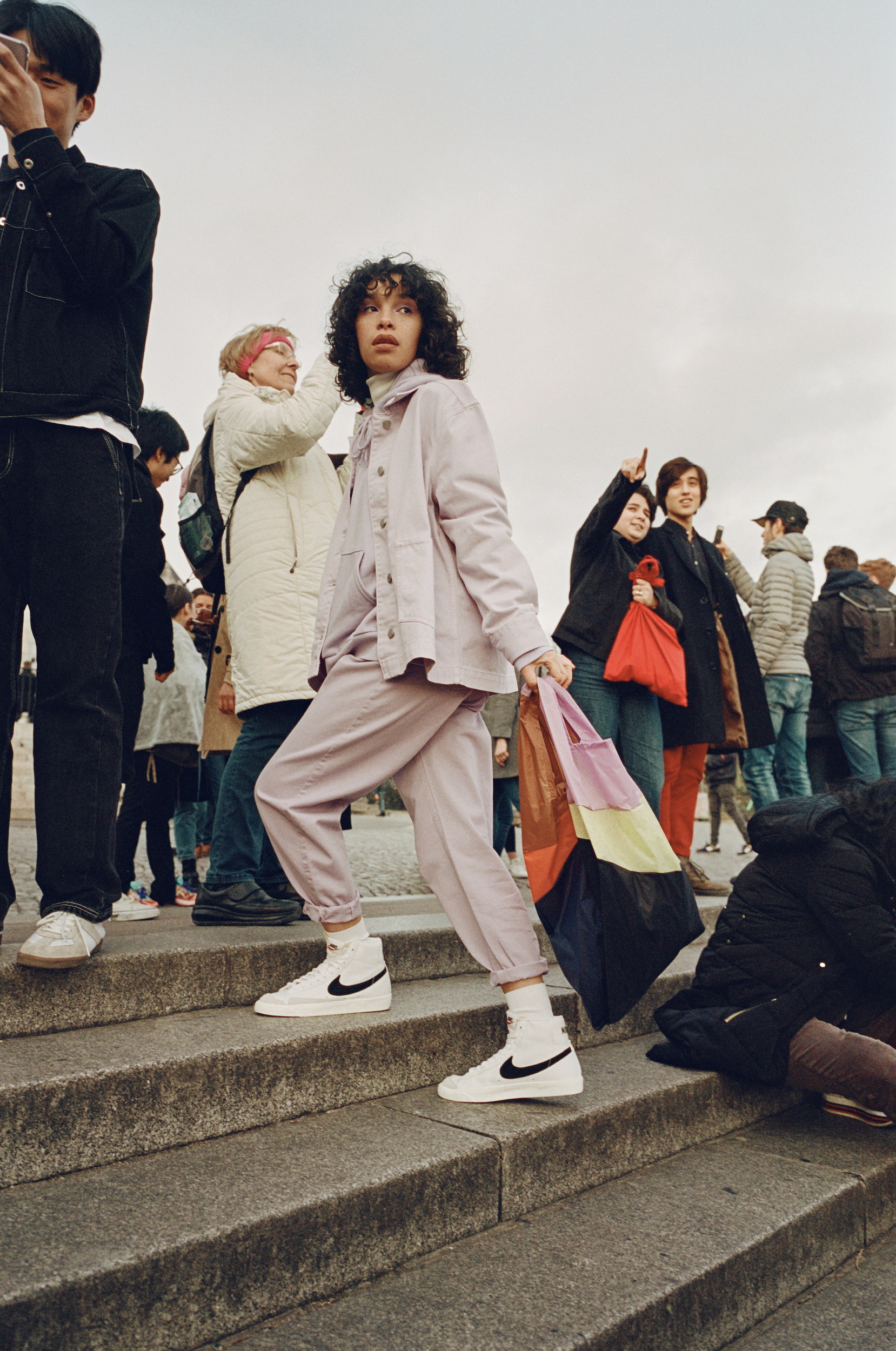 Woman in lavender pantsuit and white sneakers walking on stairs with a colorful tote bag, surrounded by blurred people.