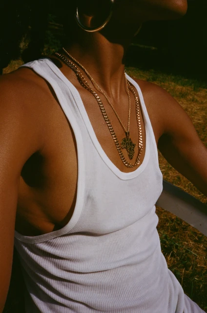 Close-up of a Black adult female wearing a white tank top, gold hoop earrings, and gold necklaces, posing outdoors in natural sunlight.