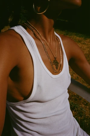 Close-up of a Black adult female wearing a white tank top, gold hoop earrings, and gold necklaces, posing outdoors in natural sunlight.