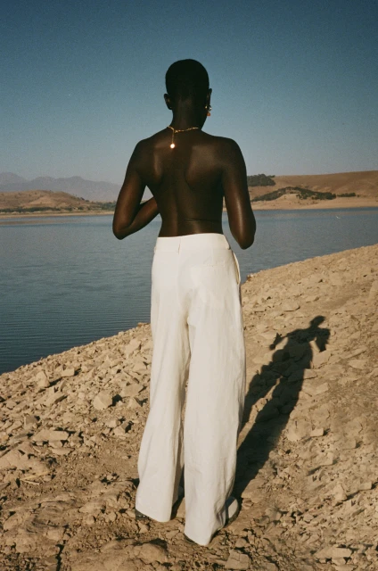 Back view of a dark-skinned person in white pants standing on a rocky beach with a blue lake and mountains in the background.