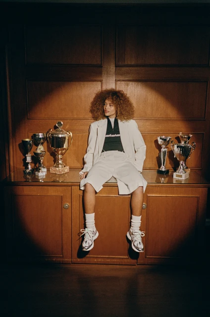 Stylish young adult with curly hair sits on a wooden cabinet with sports trophies wearing a blazer, shorts, and sneakers in a spotlight.