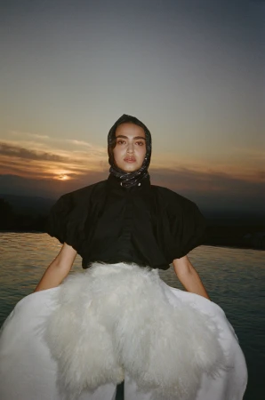 Woman wearing a dark puffed-sleeve top, headscarf, and white skirt against a sunset sky and water backdrop