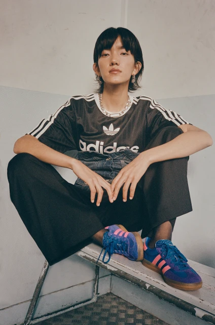 Asian female in Adidas shirt poses seated on metal structure wearing blue sneakers, black trousers, and silver jewelry in an indoor setting.
