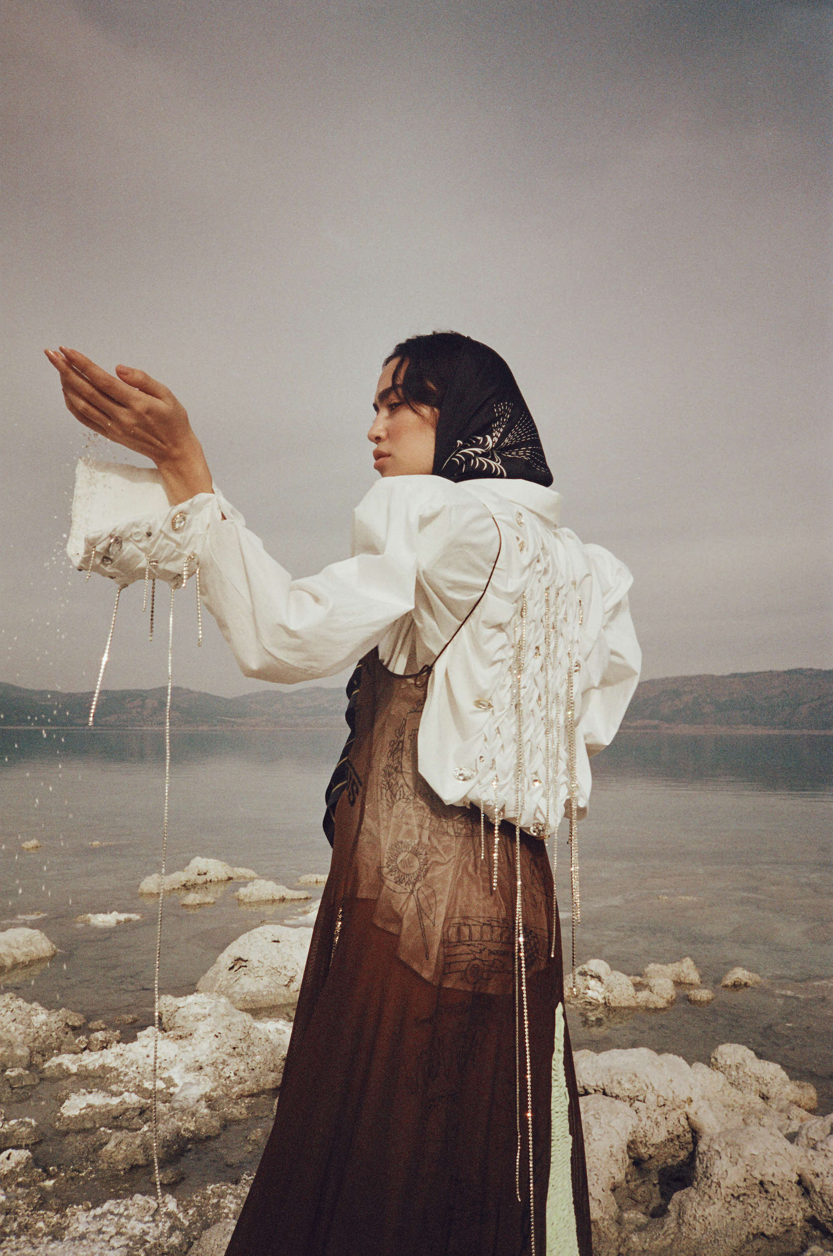 Woman in flowing brown dress, white cropped jacket, and headscarf stands near a tranquil lake, holding a white object.