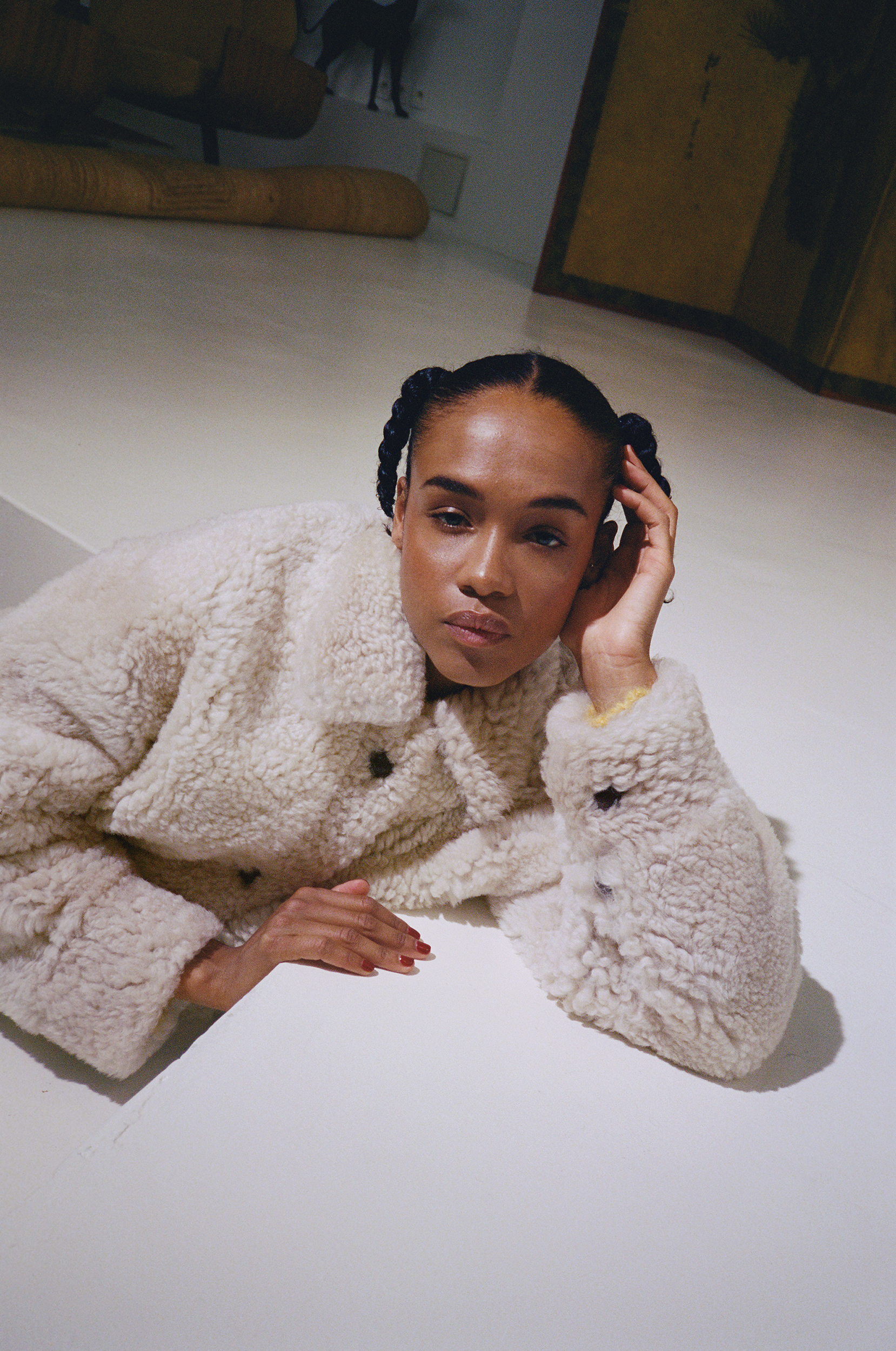 Adult female with braided pigtails poses indoors, wearing a textured shearling jacket and resting her head on her hand.