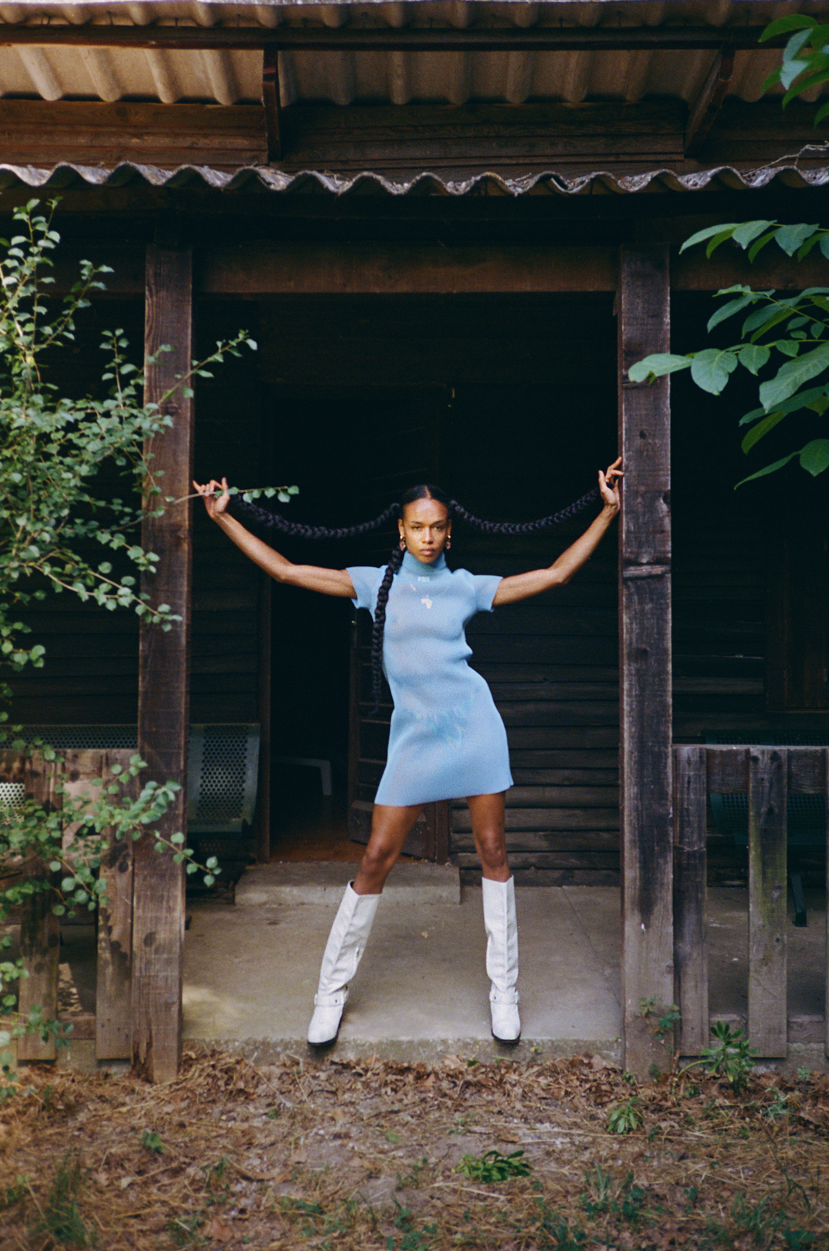 A woman in a light blue dress and white boots stands in the doorway of a weathered wooden cabin, holding her braids out to each side.