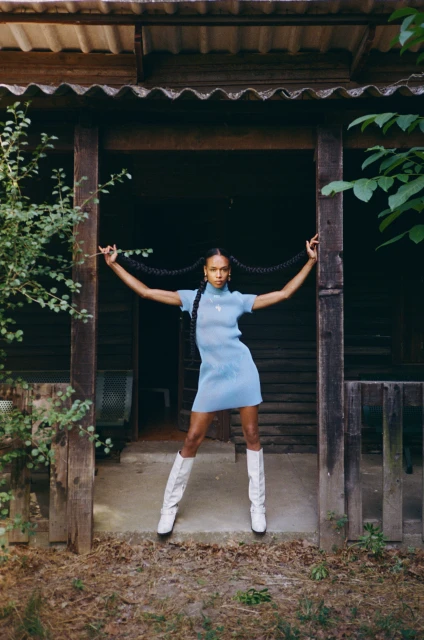 A woman in a light blue dress and white boots stands in the doorway of a weathered wooden cabin, holding her braids out to each side.